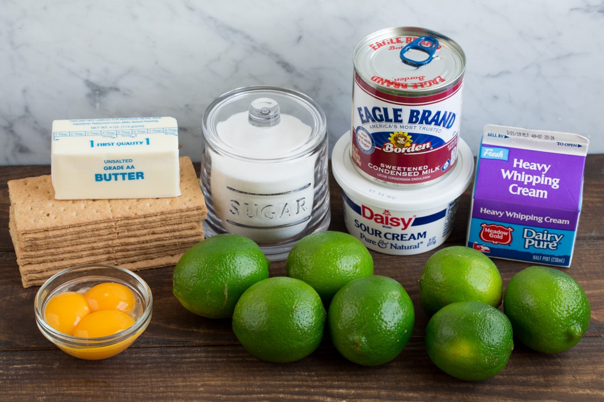 BEST EVER Key Lime Pie recipe! Eight ingredients needed for key lime pie shown here including graham crackers, butter, sugar, egg yolks, limes, sour cream, sweetened condensed milk and heavy cream.