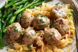 Serving of Swedish meatballs on egg noodles with a side of asparagus on a speckled cream plate.