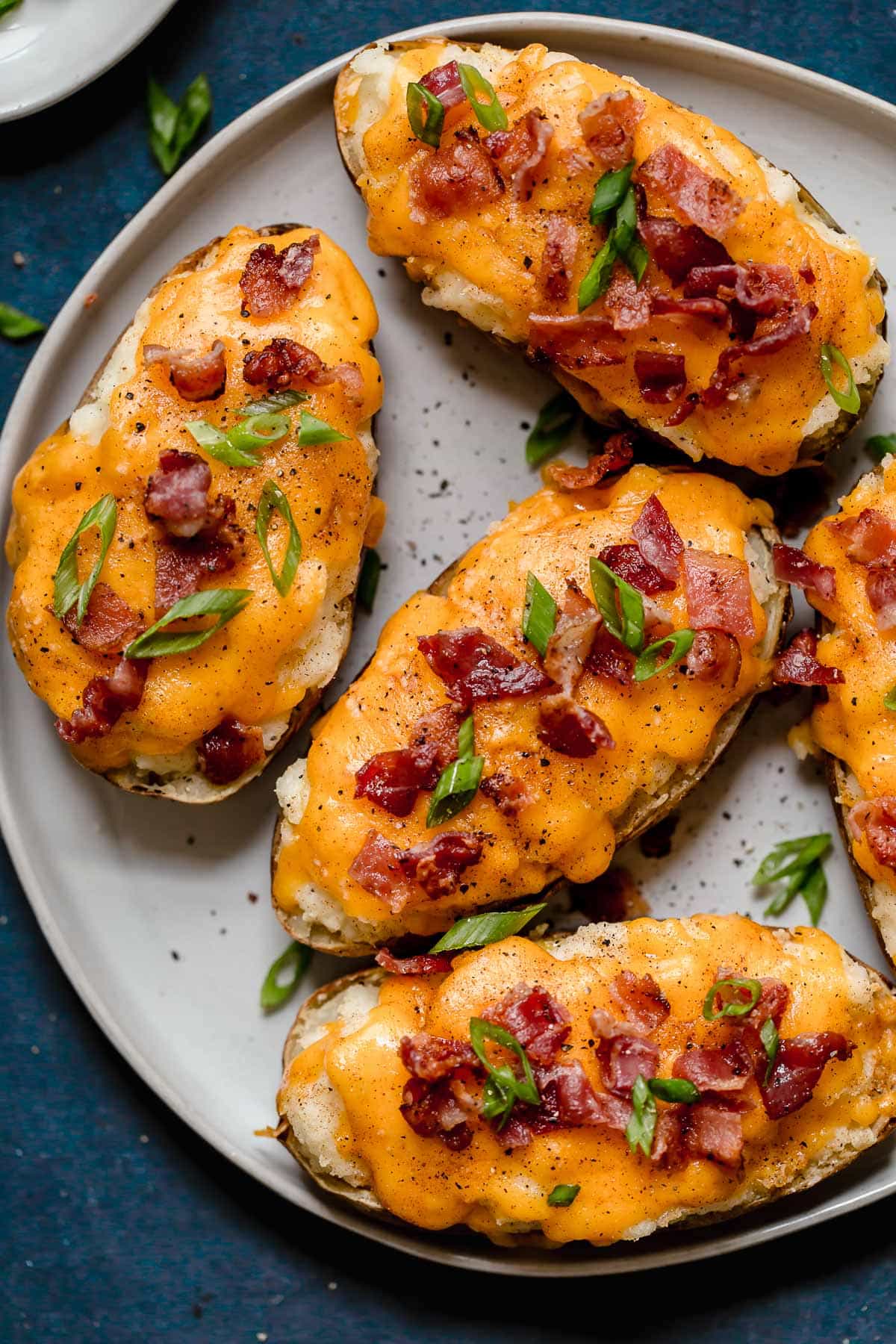 Twice Baked Potatoes Overhead image of four Twice Baked Potatoes on a white serving plate. Potato skins are filled with mashed potatoes, topped with melted cheddar cheese and garnished with bacon and green onions.