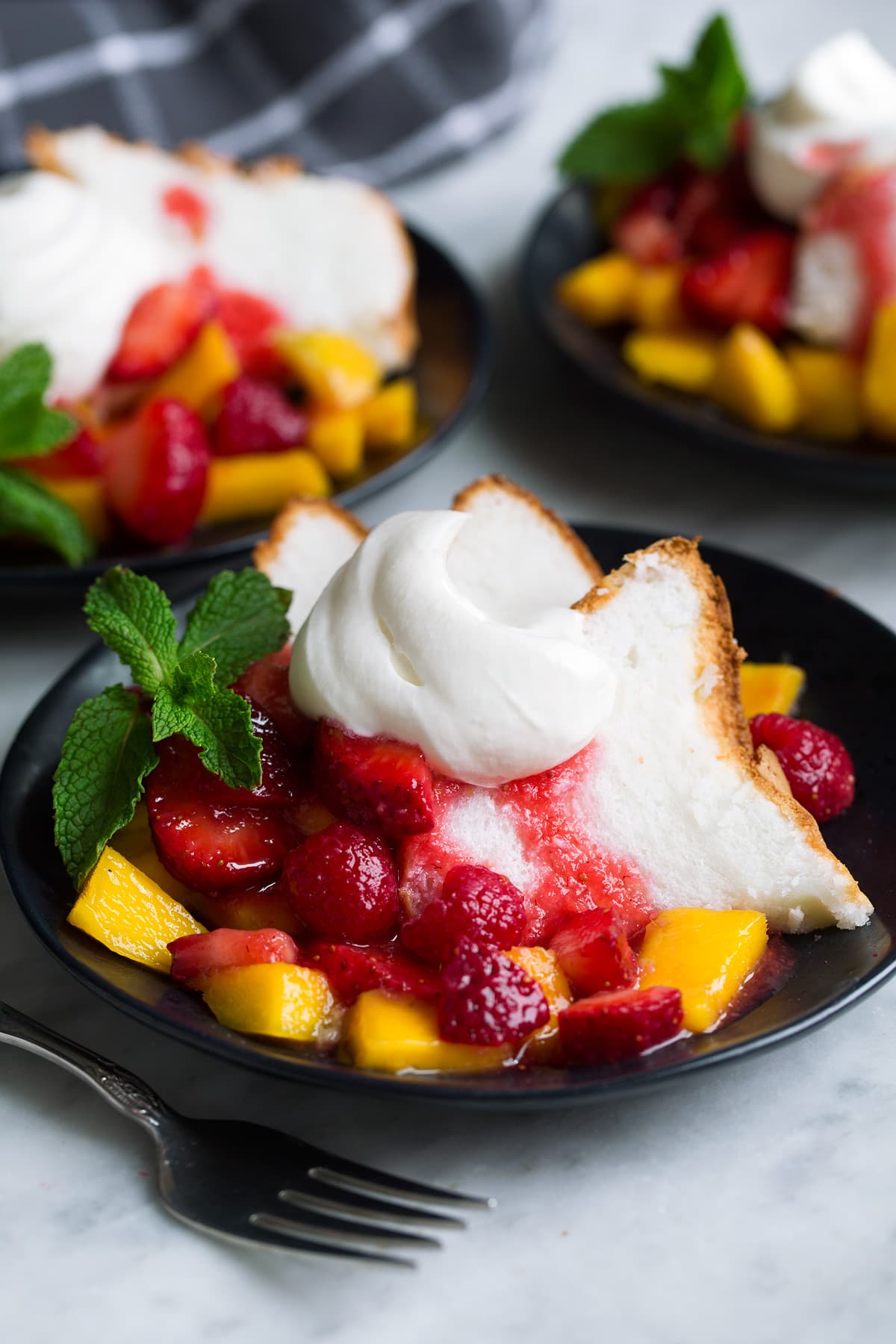 How to Make Whipped Cream Homemade whipped cream served over angel food cake with a side of fruit on a black dessert plate.
