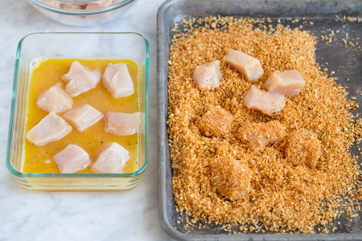 Baked Chicken Nuggets Showing how to make chicken nuggets. Tossing chicken pieces with egg then dressing in panko breadcrumb mixture on baking sheet.