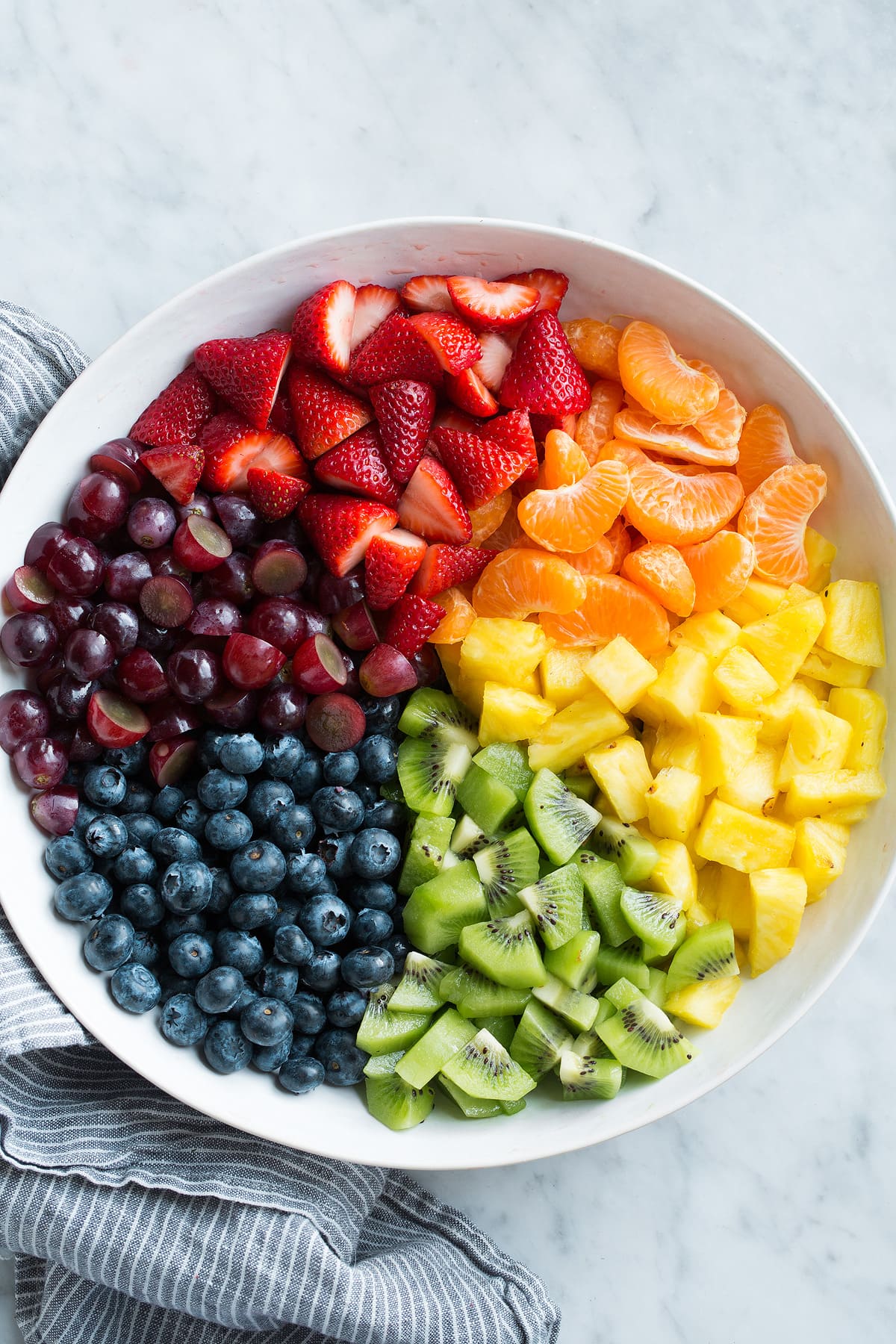 Honey Lime Rainbow Fruit Salad Fresh strawberries, mandarin oranges, pineapple, kiwi, blueberries, and grapes in sections in a large white serving bowl.