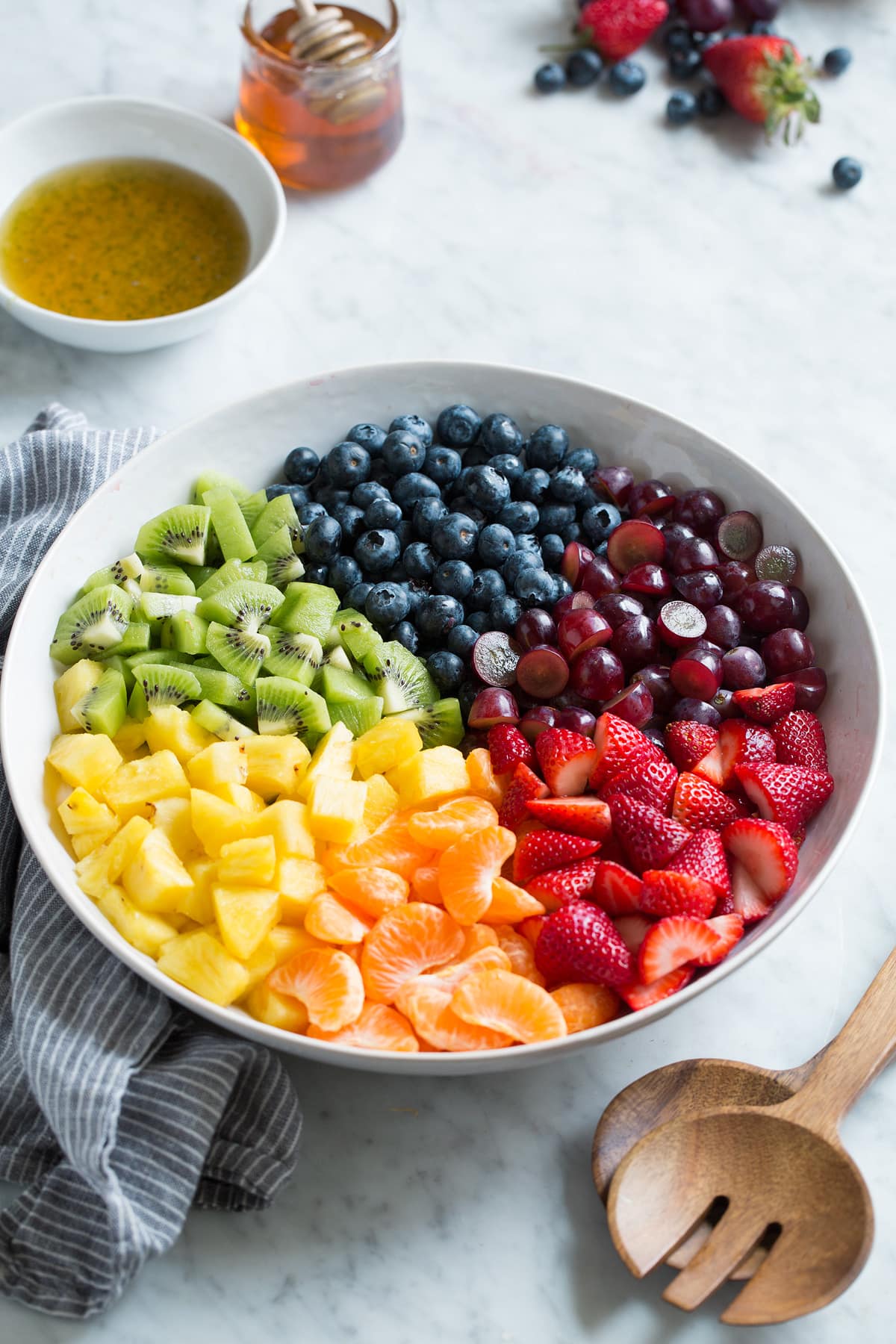 Honey Lime Rainbow Fruit Salad Fresh strawberries, mandarin oranges, pineapple, kiwi, blueberries, and grapes in sections in a large white serving bowl.