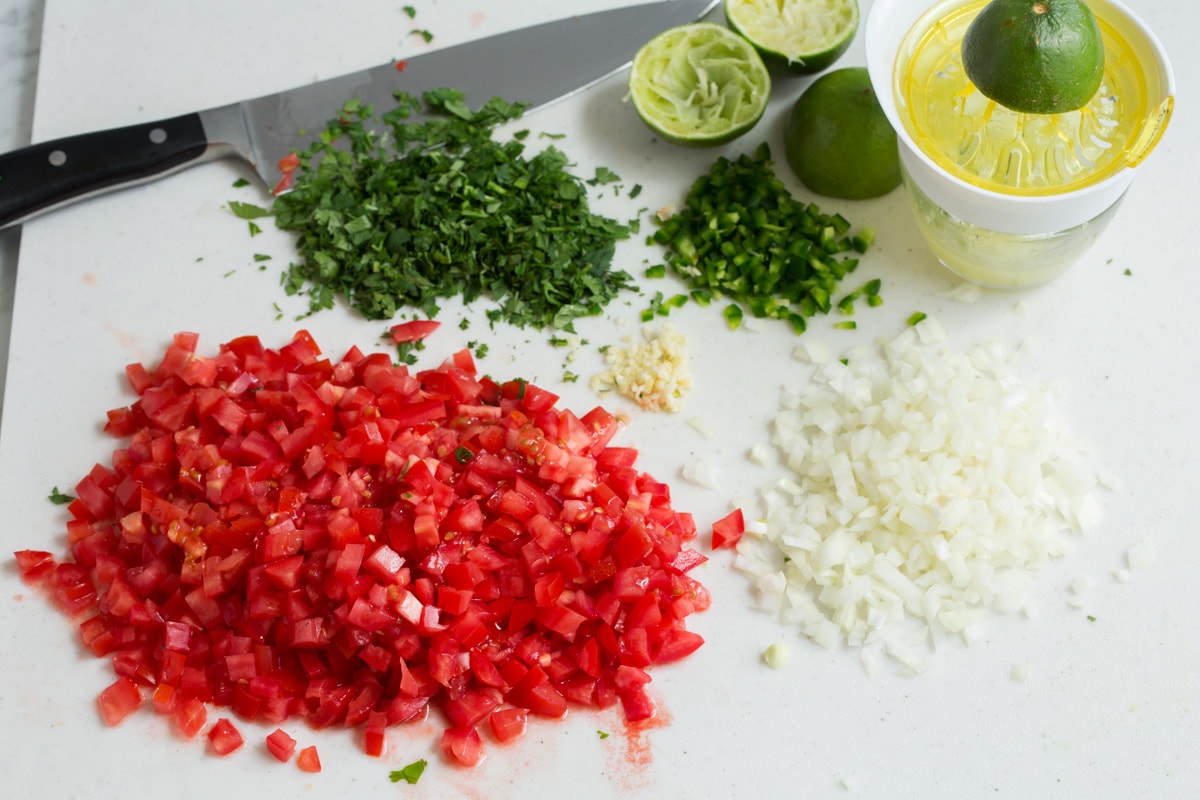 Easy Pico de Gallo! Finely chopped tomatoes, onion, cilantro, jalapeno pepper and garlic on a cutting board with a knife in the background and a juicer.