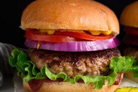 Turkey Burger layered with bun, ketchup, lettuce, turkey patty, red onion, tomato and yellow mustard. Served with a side of pickles on a white platter.