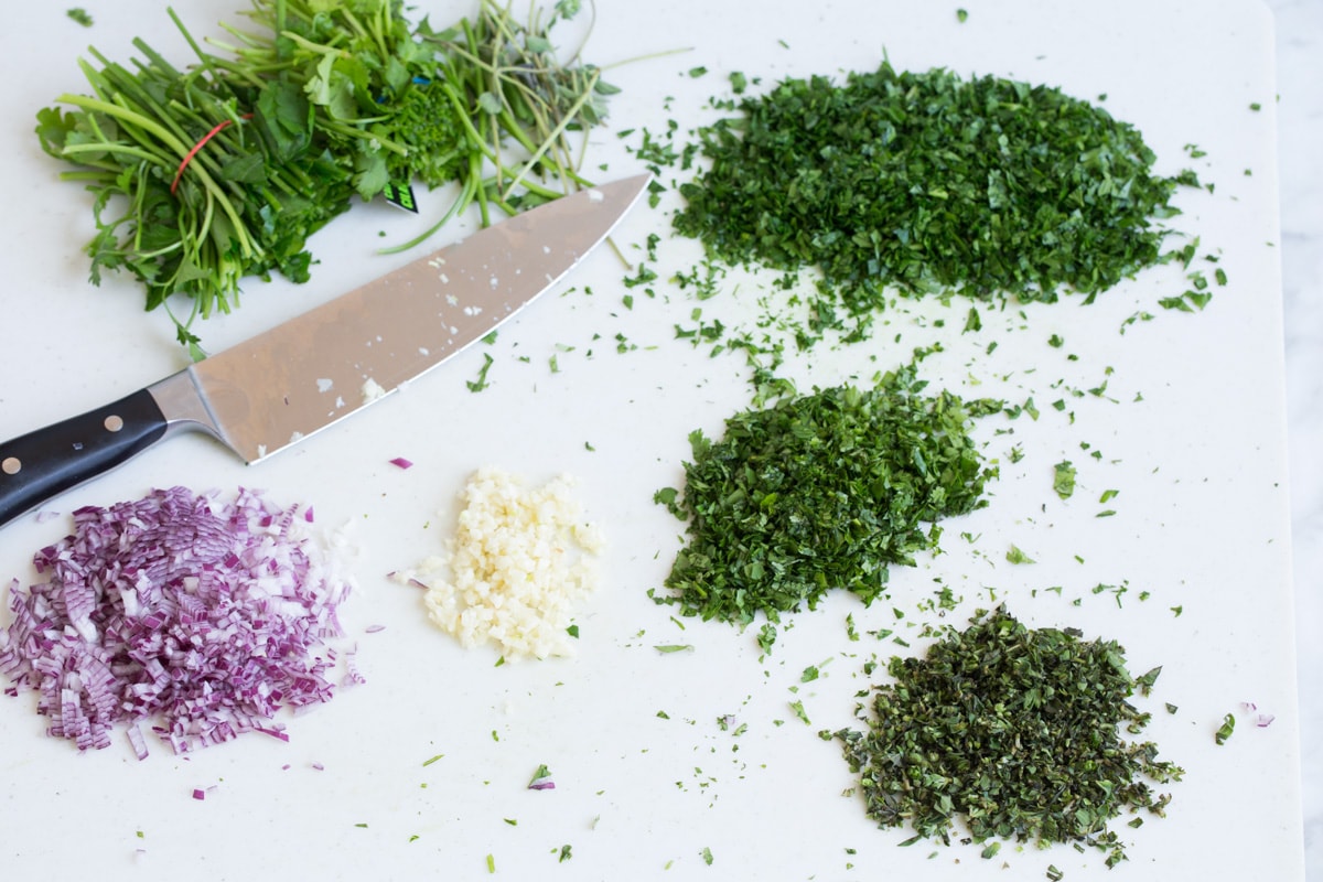 Best Chimichurri Sauce Minching up fresh herbs onion and garlic on a cutting board.