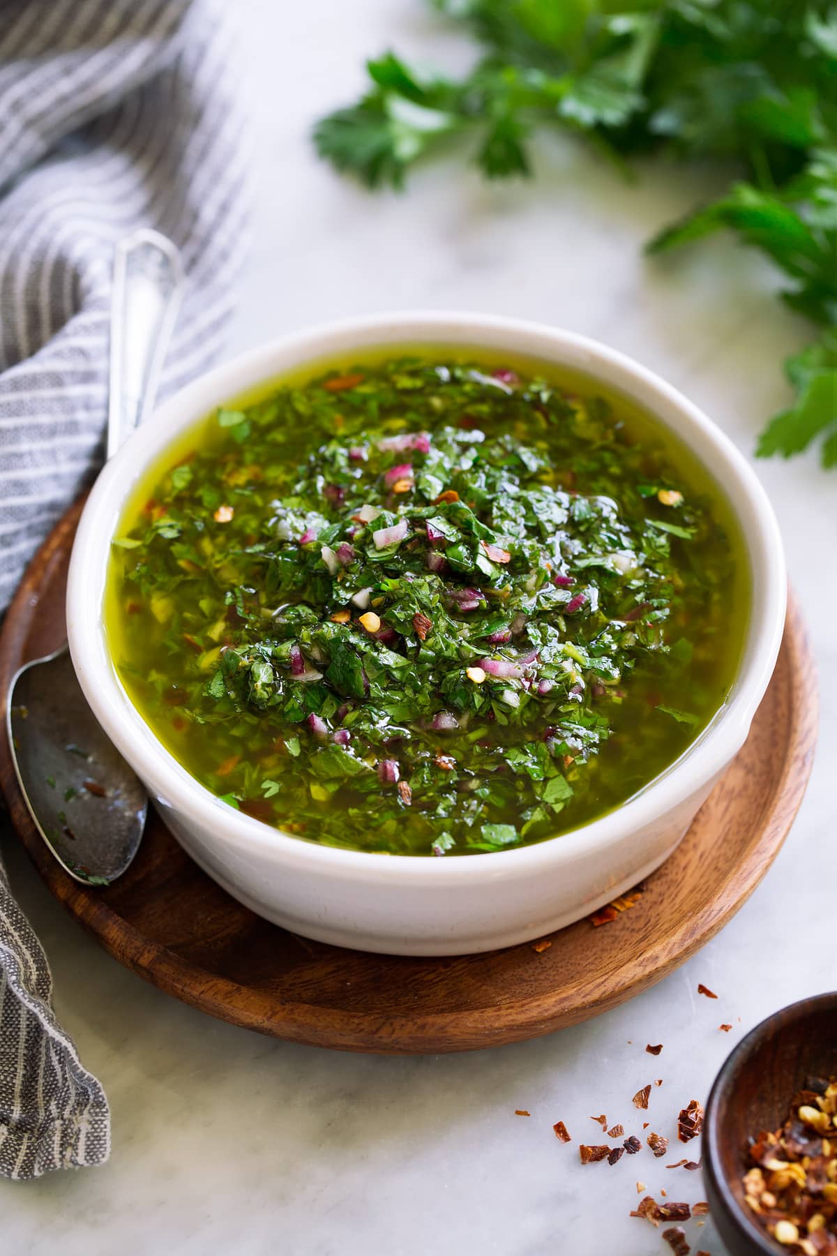 Best Chimichurri Sauce Chimichurri sauce in a small white bowl set over a wooden plate with a spoon to the side.