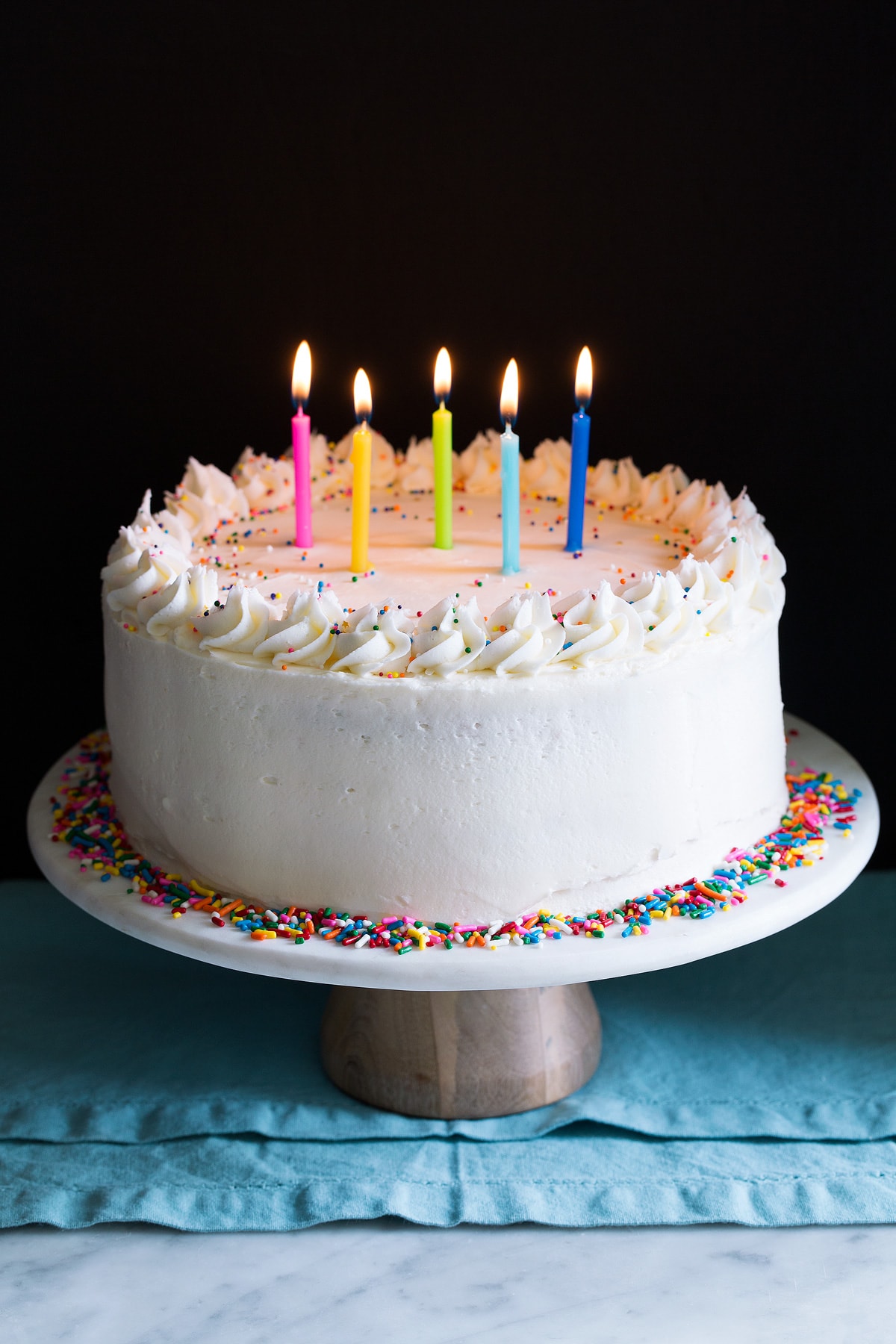 Birthday Cake - the perfect funfetti sprinkled cake! It's perfectly soft and fluffy and amazingly delicious! My kids absolute favorite cake! #cake #birthdaycake #dessert #birthday Whole birthday cake on a cake stand garnished with sprinkles and topped with lit birthday candles.