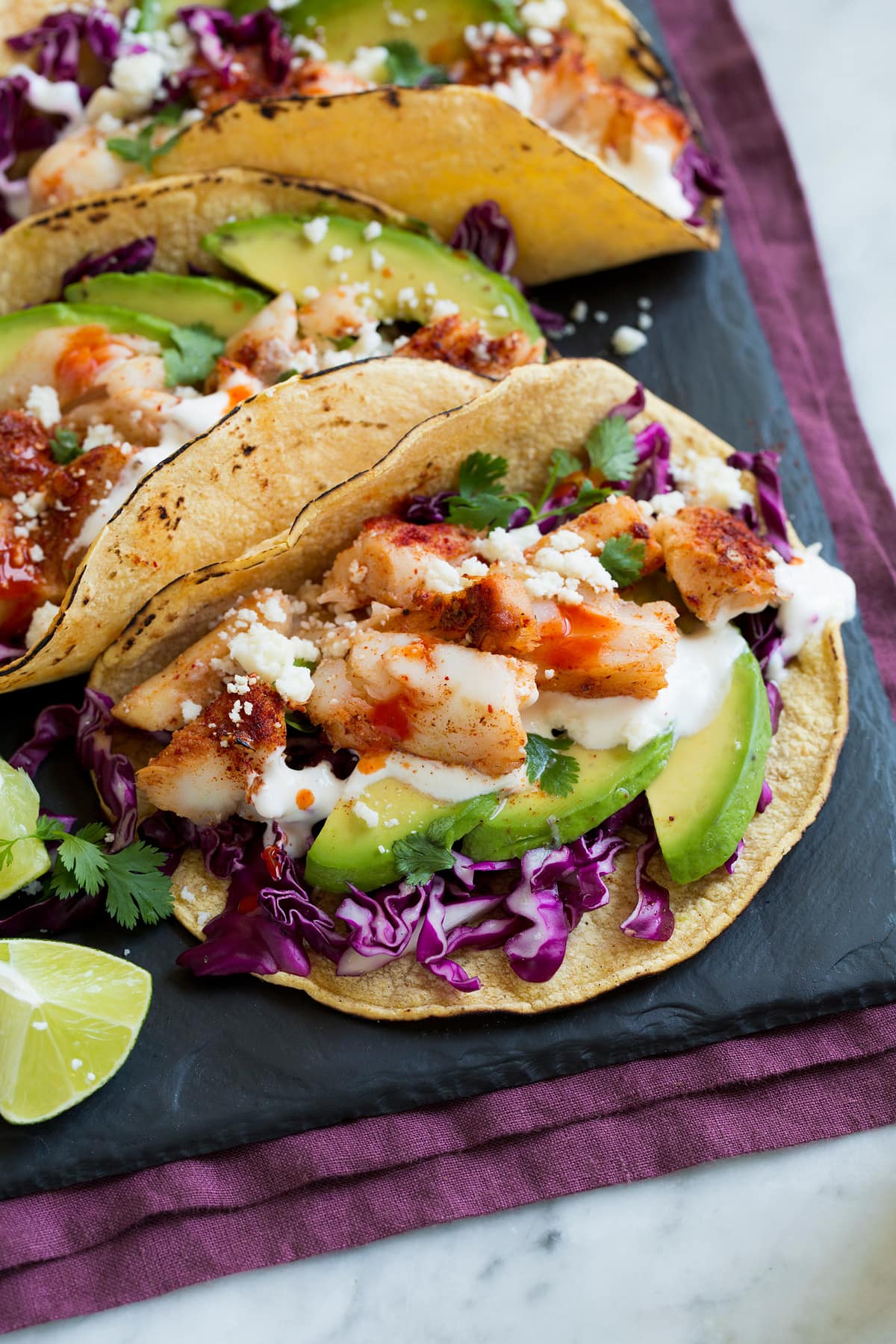 Fish Tacos - these are unbelievably delicious! Easy to make too! Includes directions for oven or grill - you'll love both, just don't omit the sauce! #fishtacos #tacos #fish #recipe Close up image of fish tacos layered with purple cabbage, avocado, fish taco sauce, cod, talapia, and cotija.