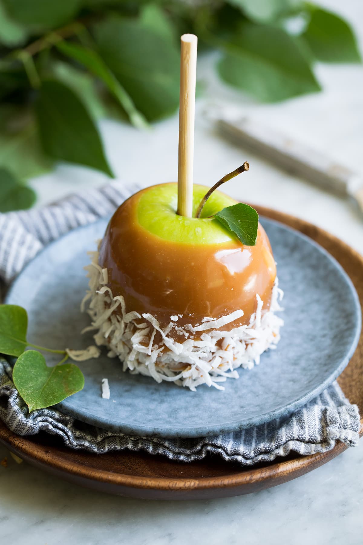 Easy Caramel Apples Single caramel apple rolled in shredded coconut sitting on a blue plate on top of a wooden plate.