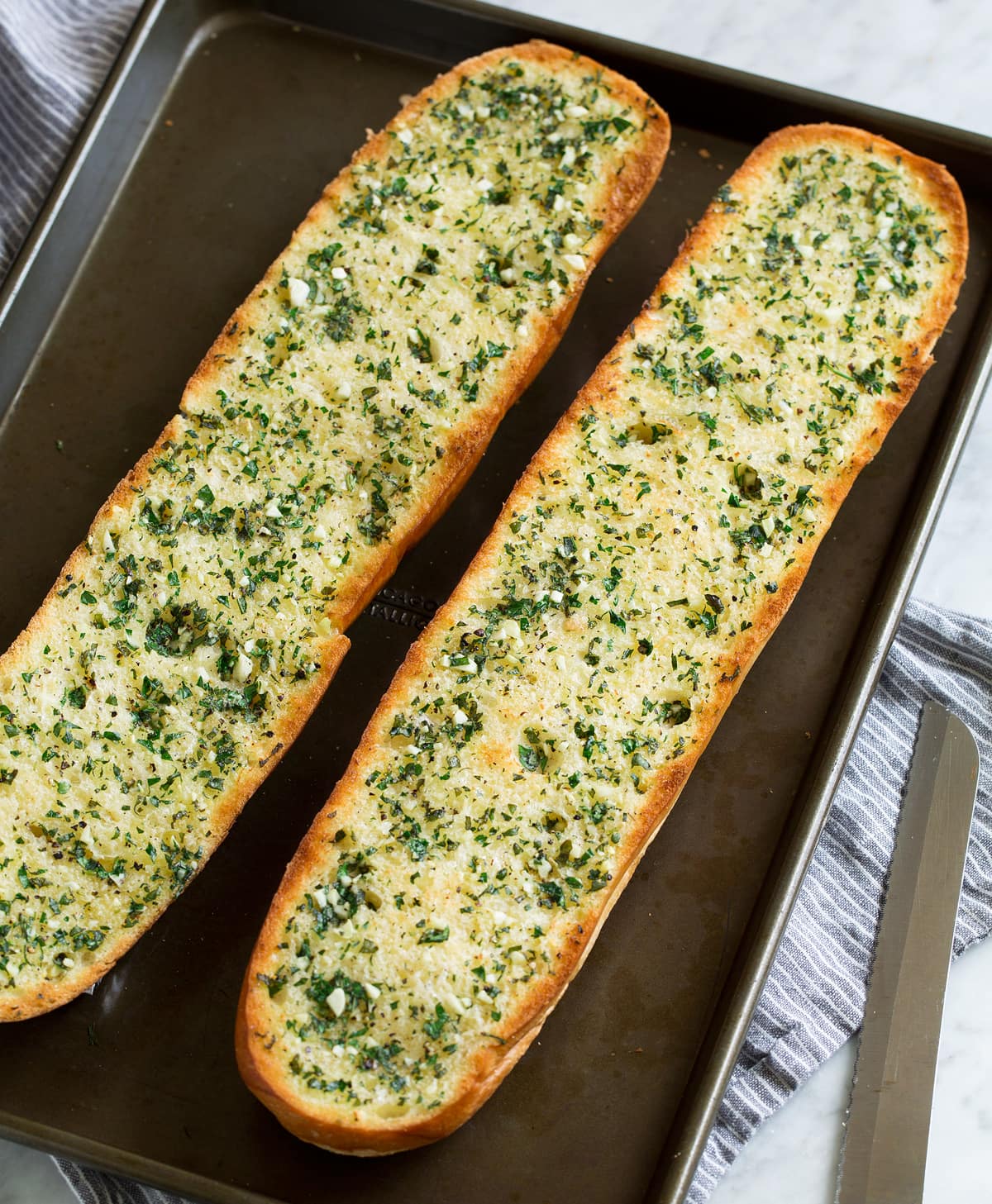 Garlic Bread - super fast, super easy and incredibly flavorful! Skip the margarine coated garlic bread from the store and make this butter version instead, it's a hundred times better! #garlicbread #sidedish #bread #appetizer Two garlic bread halves on a dark baking sheet. Shown after toasting.