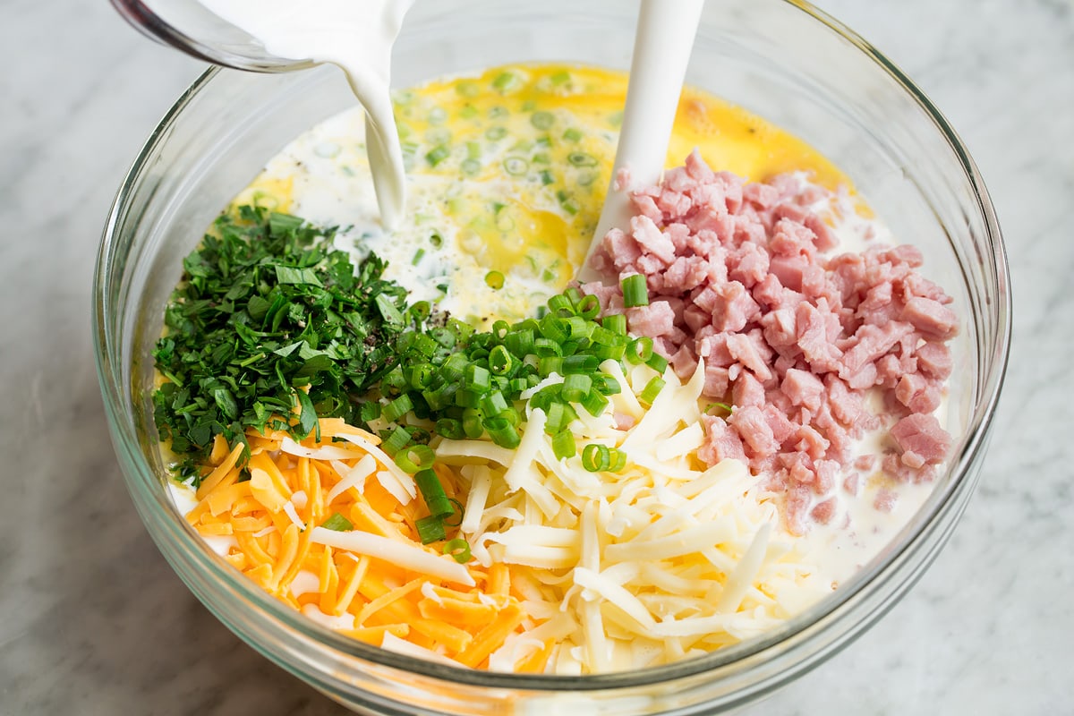 Breakfast Casserole Pouring half and half into glass bowl filled with eggs, ham, shredded cheese, green onions and parsley.