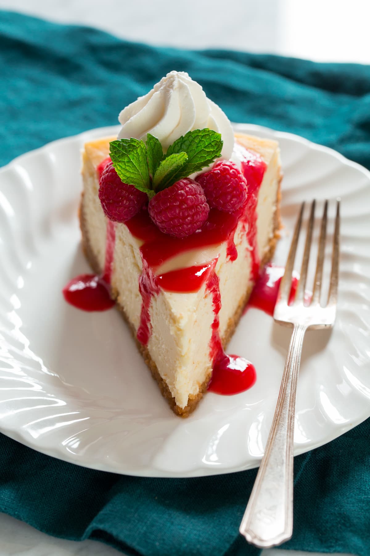 The BEST Cheesecake Recipe! Slice of cheesecake topped with raspberry sauce, fresh raspberries, whipped cream and mint.