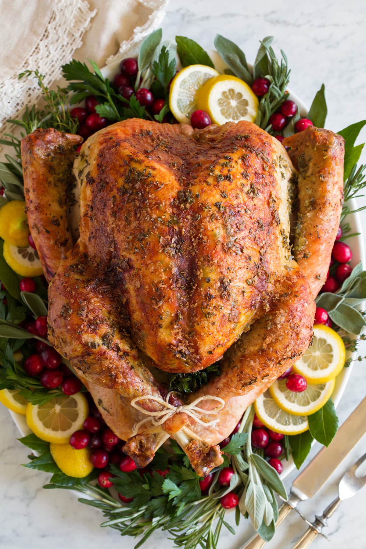 Roast Turkey - easy to follow recipe that turns out tender, juicy and full of flavor! A holiday staple! #turkey #thanksigiving Overhead image of whole turkey on a white platter garnished with fresh herbs, lemon slices and cranberries around it.