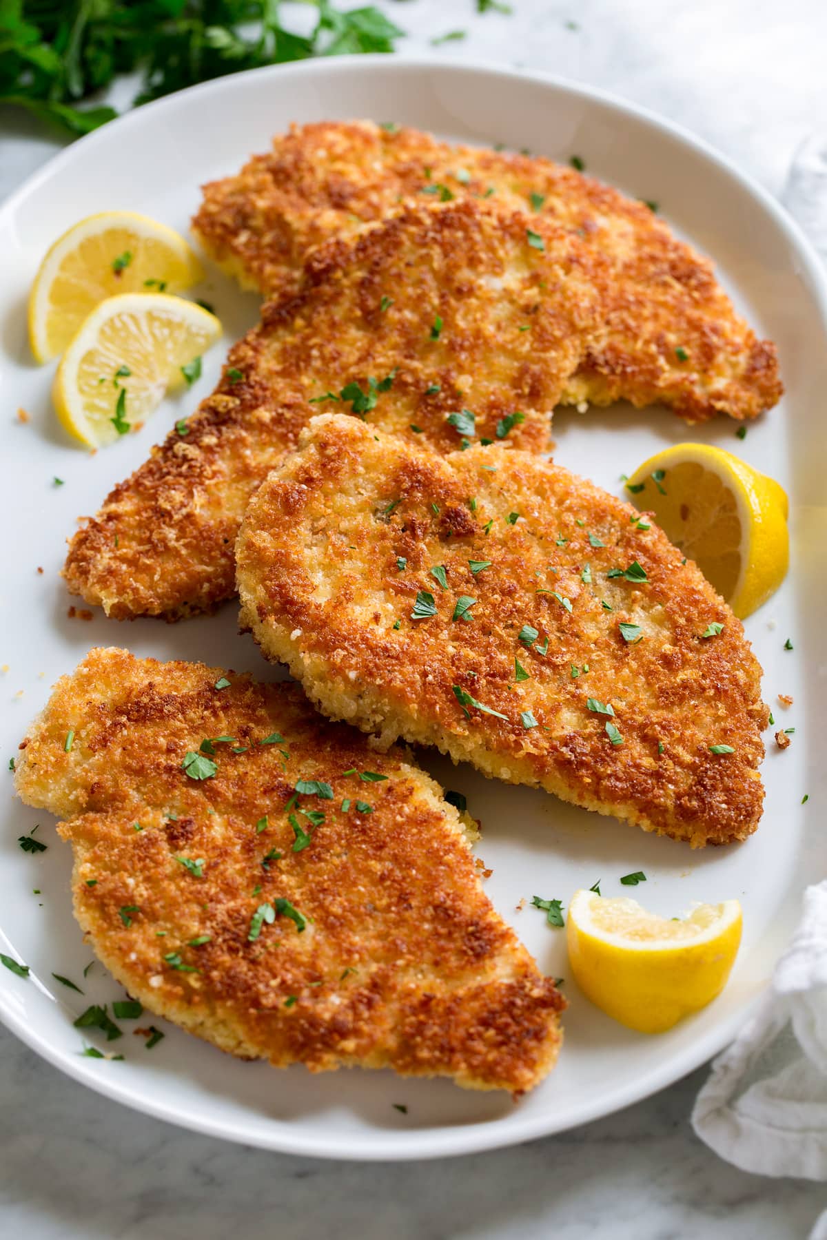 Parmesan Crusted Chicken Parmesan Crusted Chicken cutlets shown on a white serving platter from a side angle. Lemons are shown to the side and parsley is garnished over.