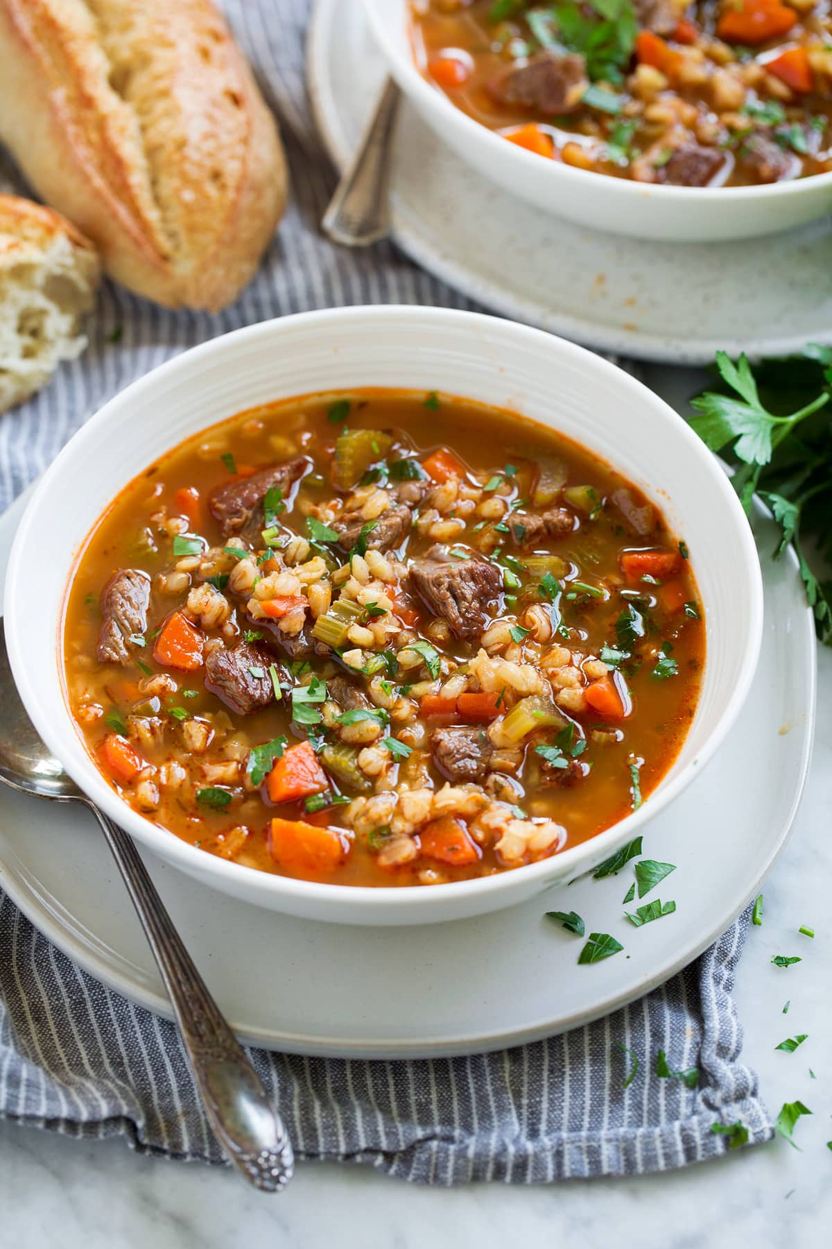 Beef and Barley Soup White bowl set over a white plate filled with beef and barley soup with a second serving shown in the background. Also shown is a side serving suggestion of fresh bread.