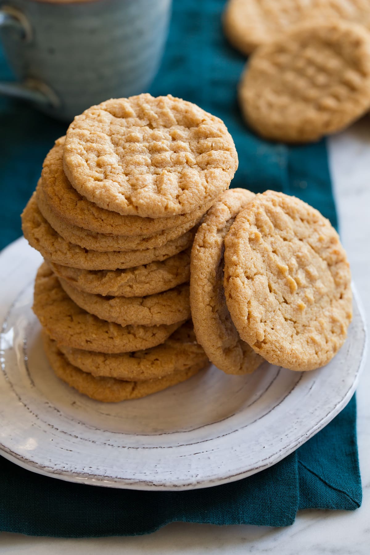 3-Ingredient Peanut Butter Cookies Three ingredient peanut butter cookies stacked on a white plate with a pile of cookies and mug in the background.