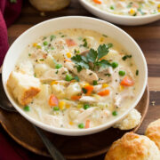 Two bowls of chicken pot pie soup in white serving dishes set over wooden a wooden tabletop. Biscuits are served to the side.