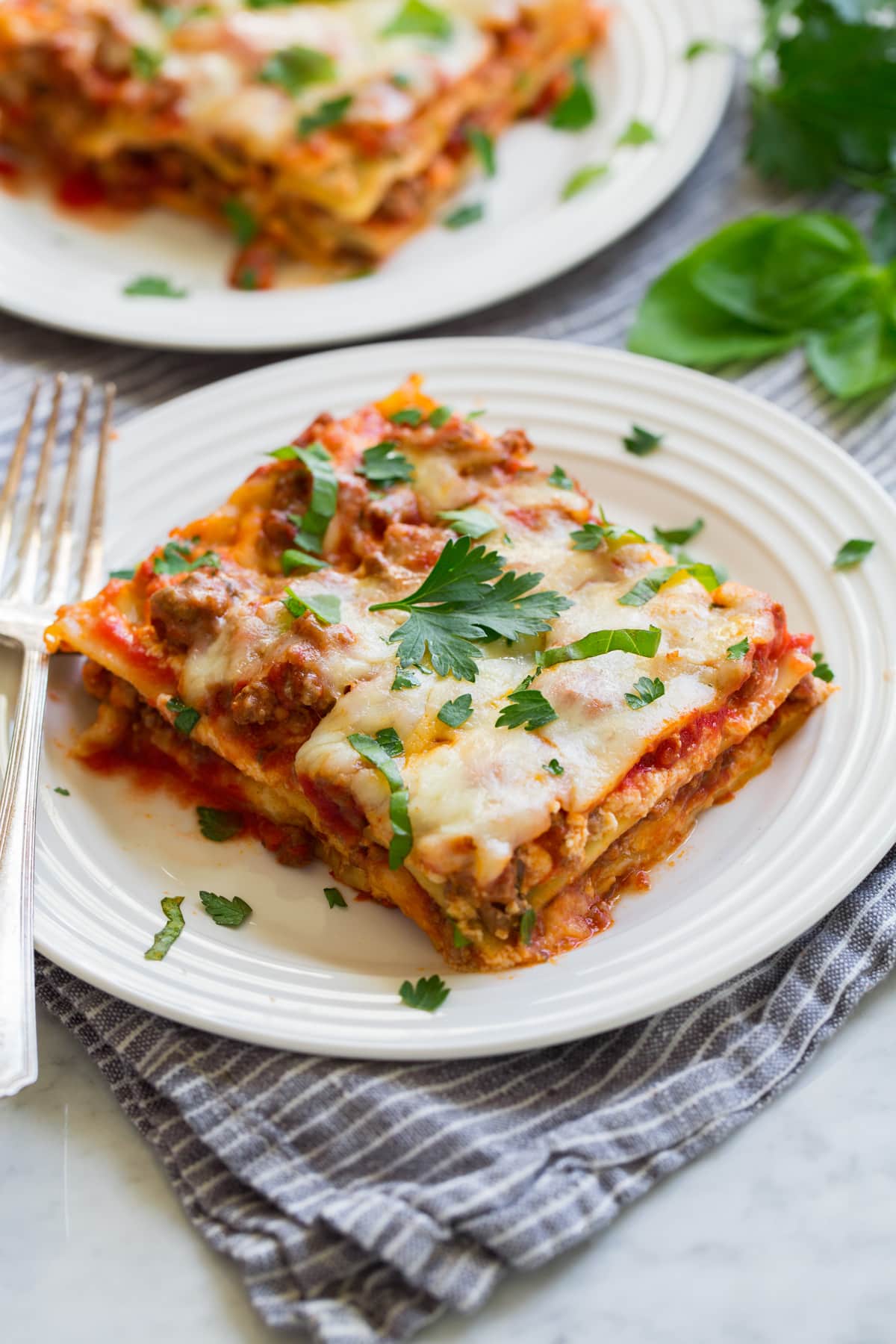 Easy Lasagna Recipe Slice of easy lasagna on a white serving plate with a second serving shown in the background. Plates are sitting on a grey striped napkin on a marble surface.