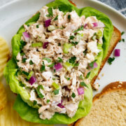 Tuna salad atop bread slices and lettuce leaves. Shown on a white plate with a side of potato chips.