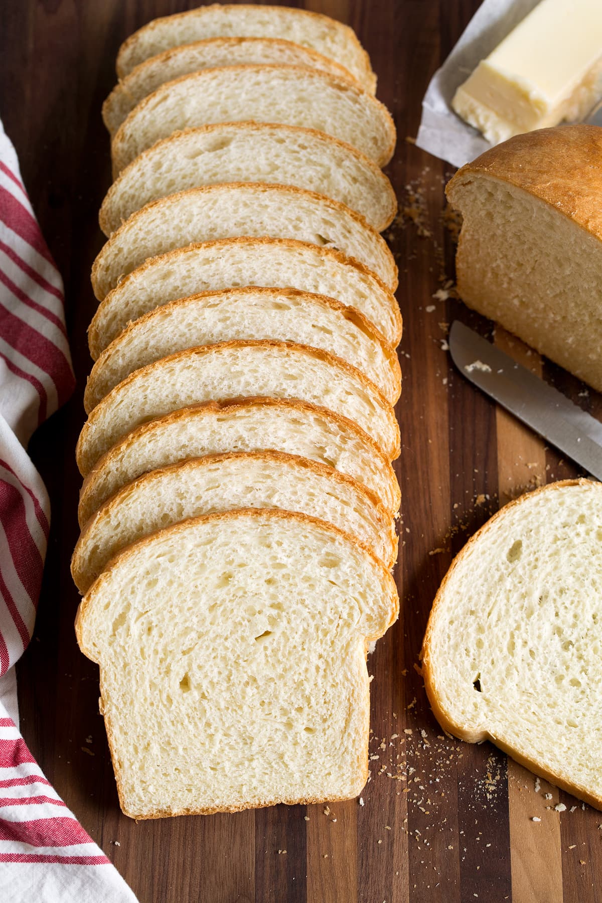 Homemade White Bread Recipe Row of fresh fluffy white bread on a cutting board with a kitchen cloth to one side and a knife and butter on the other side.