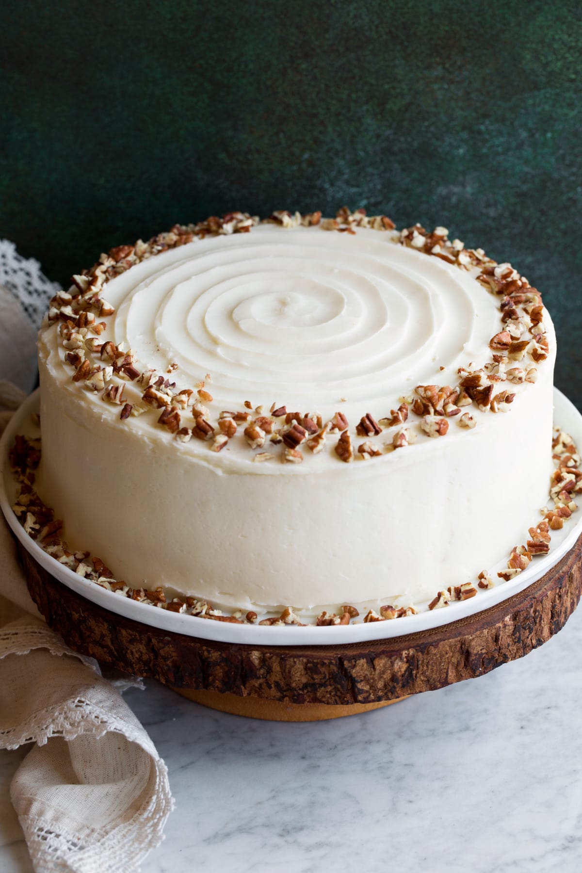 Cream Cheese Frosting Cake on a wooden cake stand covered in cream cheese frosting and garnished with a wring of chopped pecans on top.