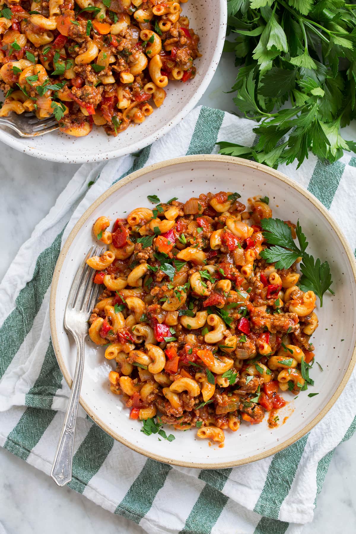 Goulash Serving of ground beef and macaroni goulash shown overhead in a white pasta bowl set over a green and white stripped napkin with parsley to the side.
