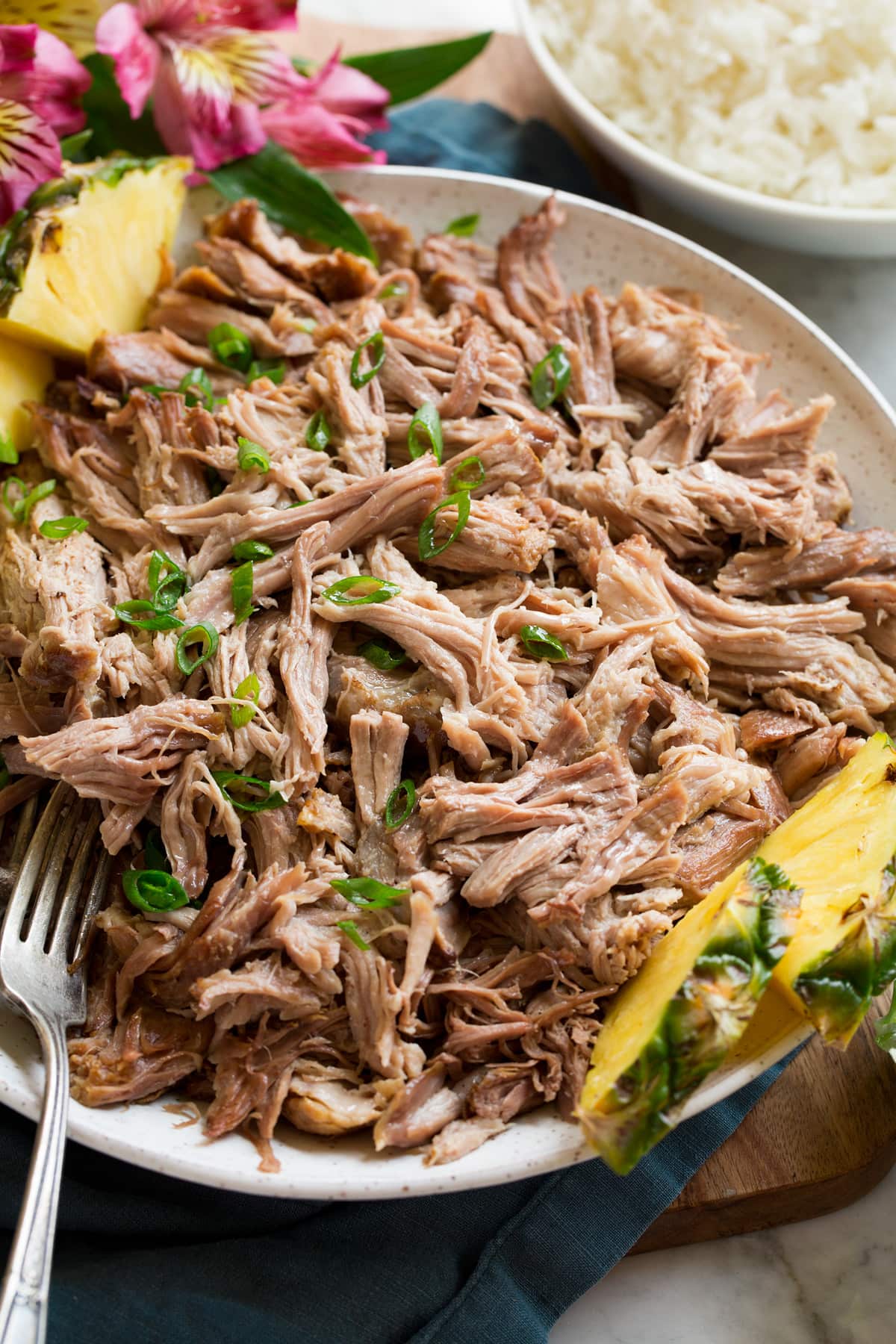 Kalua Pork Shredded kalua pork shown from the side in a white serving bowl set over a blue napkin and wooden platter. Pineapple slices and tropical flowers are shown to the side.