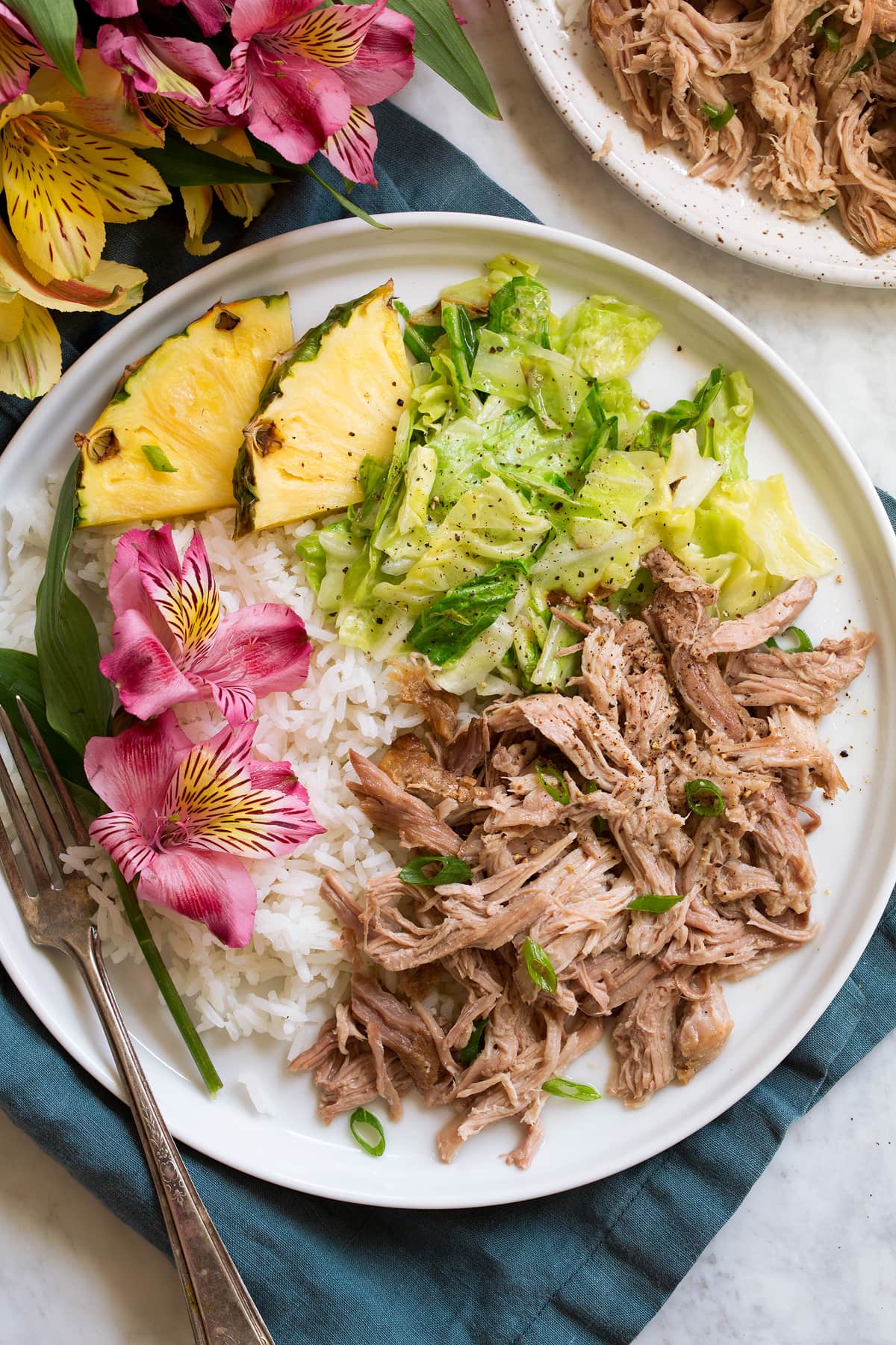 Kalua Pork Single serving of shredded kalua pork on a white serving plate shown with sides of white rice, sautéed cabbage, fresh pineapple and flowers.