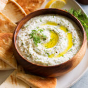 Bowl of homemade tzatziki sauce in wooden bowl on a platter with pita bread to the side.
