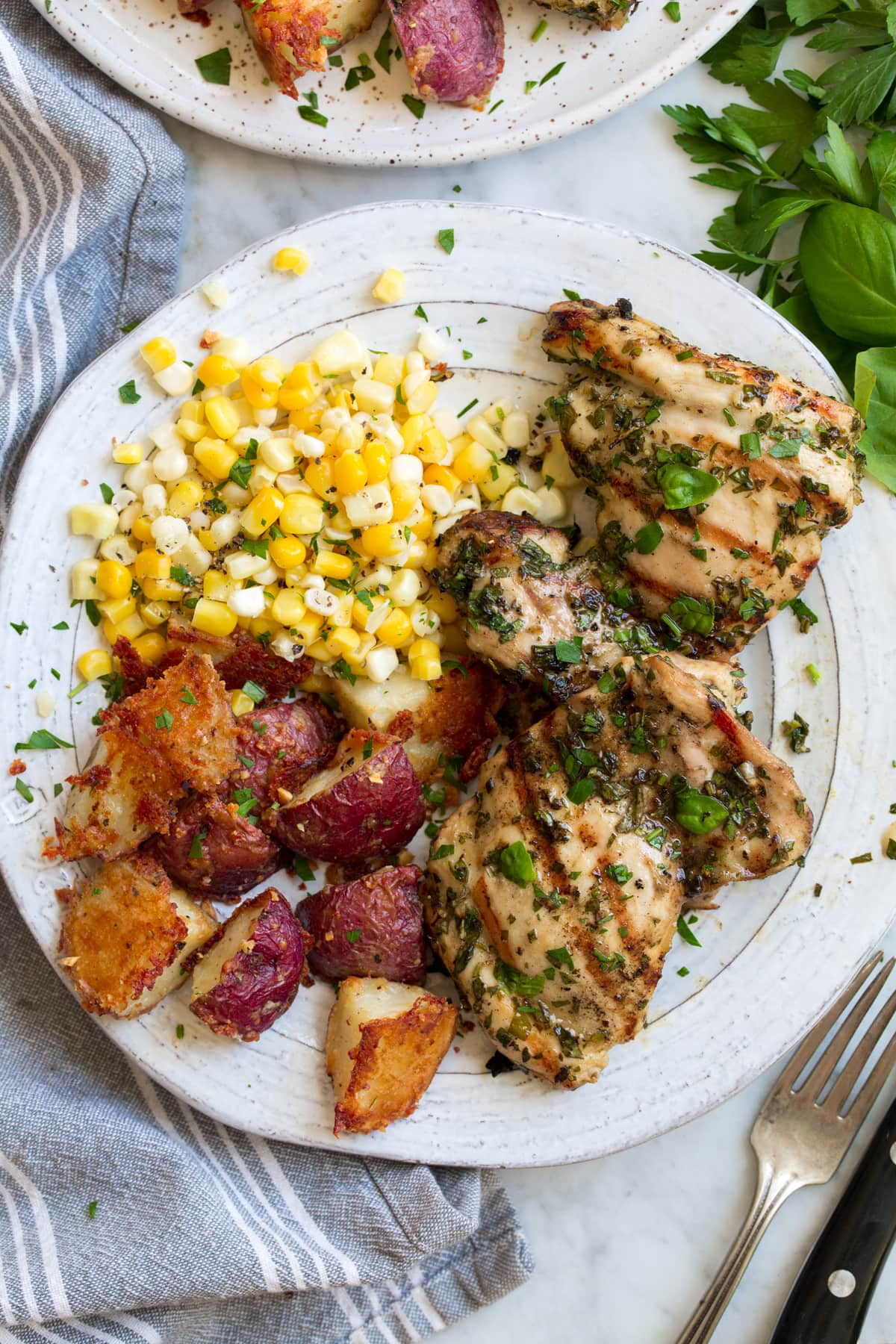 Garlic Herb Grilled Chicken Overhead image of garlic and herb marinated grilled chicken thighs with corn and roasted red potatoes as sides. Food is resting on a white plate set over a marble surface.