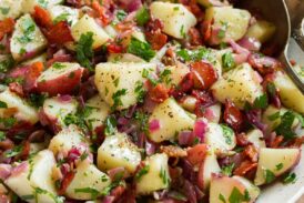 German potato salad shown in a white bowl. A silver serving spoon and fork are resting to the side, bowl is sitting on a wooden board with parsley in the background.