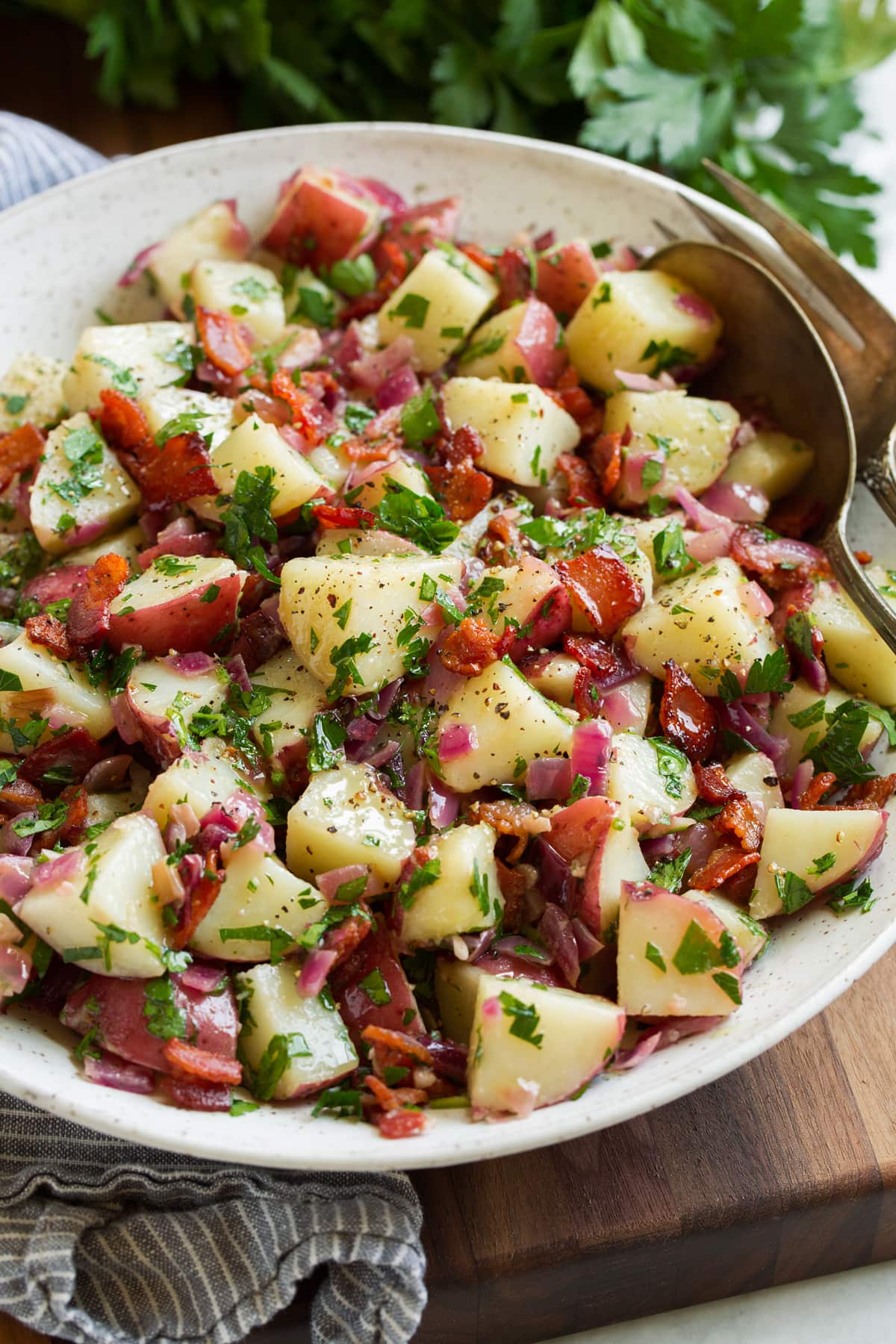 German potato salad shown in a white bowl. A silver serving spoon and fork are resting to the side, bowl is sitting on a wooden board with parsley in the background.