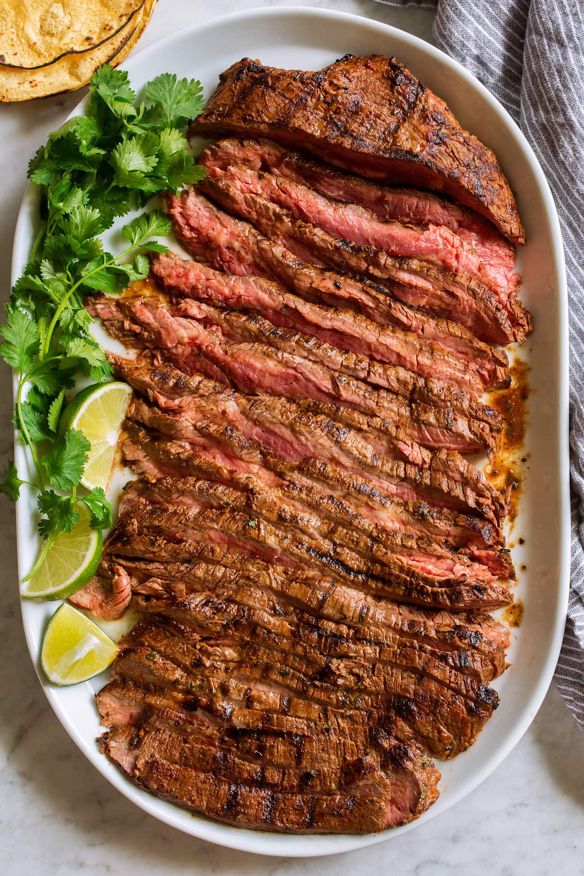 Carne Asada Overhead image of sliced carne asada on an oval white serving platter with cilantro, limes and tortillas on the side.