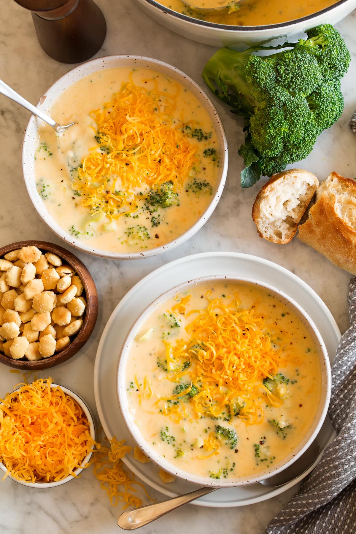 Broccoli Cheese Soup Image overhead of two servings of broccoli cheese soup in serving bowls. Crackers, broccoli, bread and cheese are shown to the side.