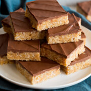 Pyramidal shaped stack of scotcheroos on a white plate, plate is set over a blue cloth and a wooden platter on a marble surface.