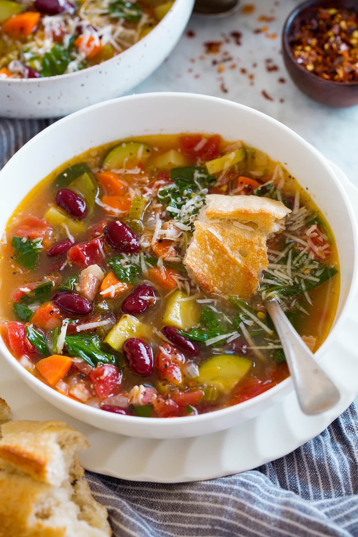 Image of serving of minestrone soup in a white bowl set over white plate. Soup is garnished with parmesan and topped with bread.