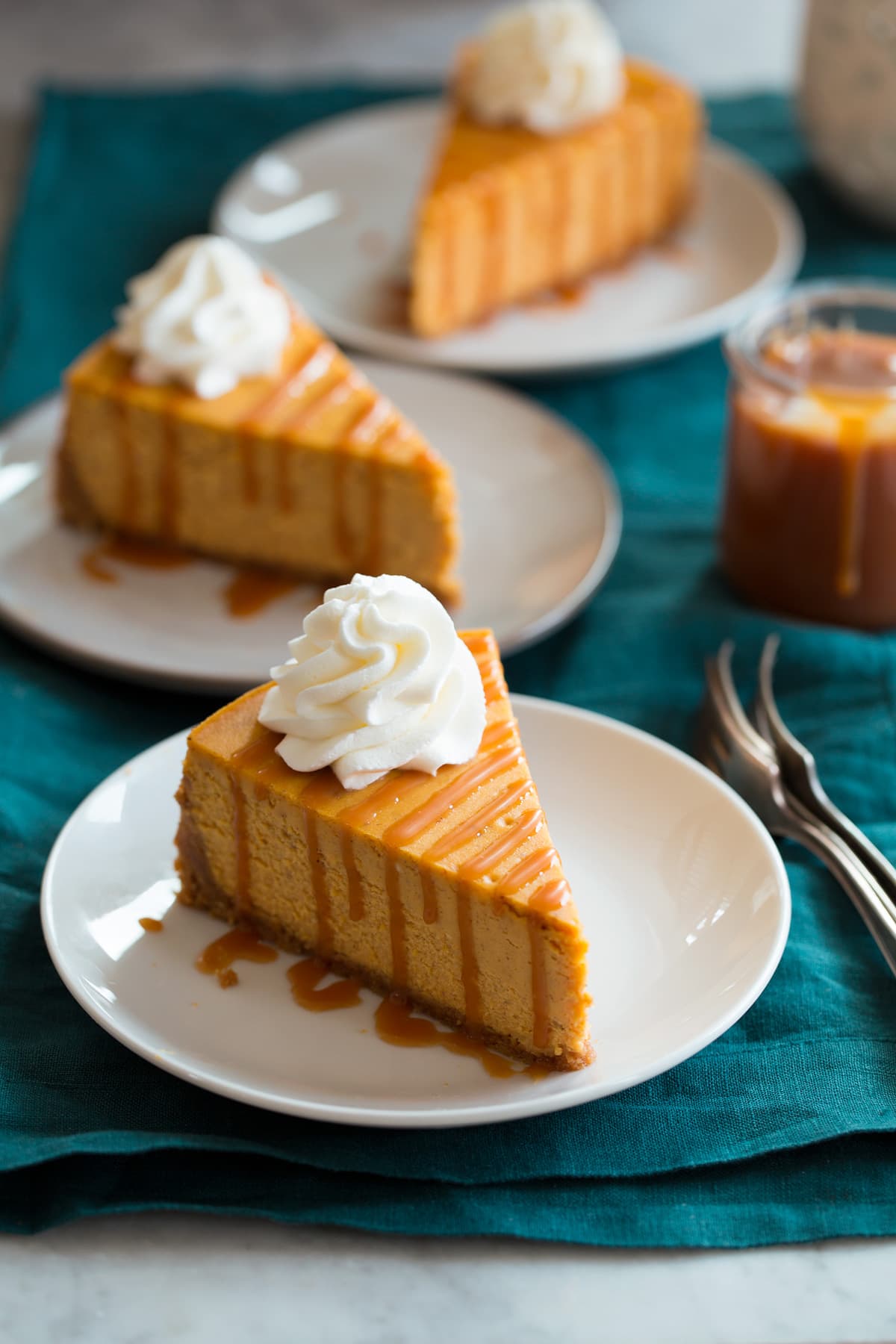 Pumpkin Cheesecake Three slices of pumpkin cheesecake shown on small white dessert plates set over a teal colored cloth.