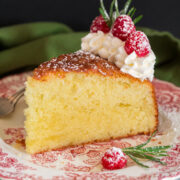 Photo of slice of olive oil cake decorated with whipped cream, raspberries and rosemary. Shown on a decorative red plate.