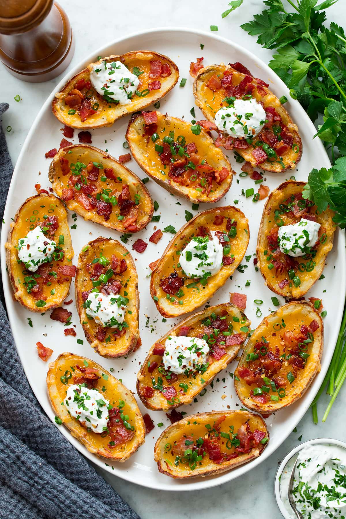 Potato Skins Potato skins on a white oval serving platter shown from above. Potato skins are garnished with green onions, bacon and topped with a dollop of herbed sour cream.