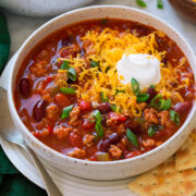 Serving of turkey chili in a white bowl set over a white plate with a spoon and saltine crackers to the side. Chili is topped with shredded cheddar cheese and sour cream.