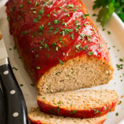 Photo of turkey meatloaf on a white oval serving platter. Parsley is sprinkled over and whole parsley is to the side. A large knife is shown to the side.