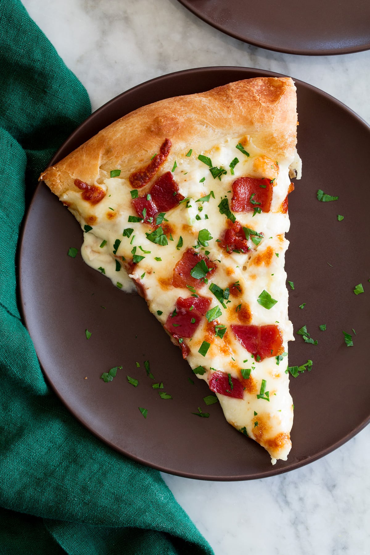 Chicken Alfredo Pizza Slice of chicken Alfredo pizza topped with bacon set on a brown plate over a marble surface.