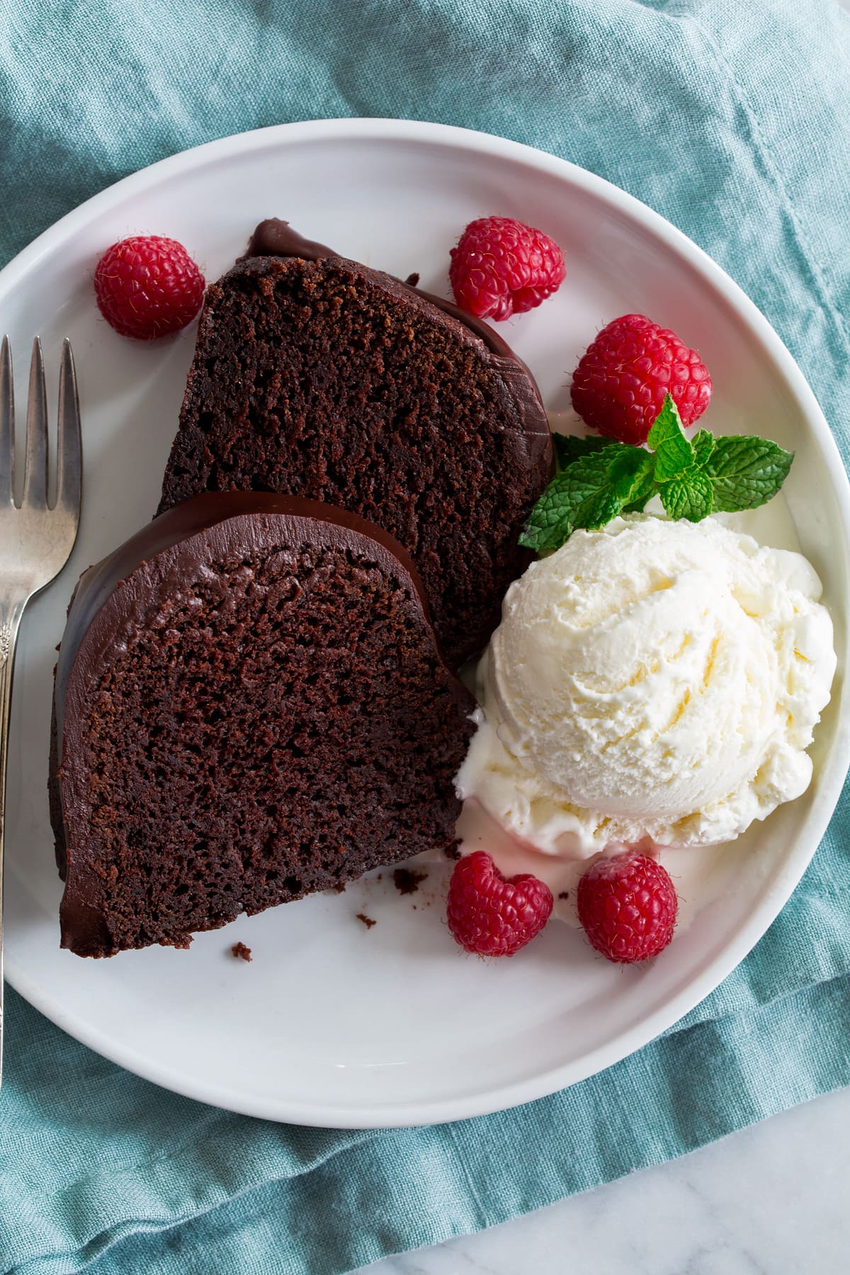 Chocolate Bundt Cake Two slices of chocolate bundt cake on a white plate with a scoop of vanilla ice cream, raspberries and mint to the side.