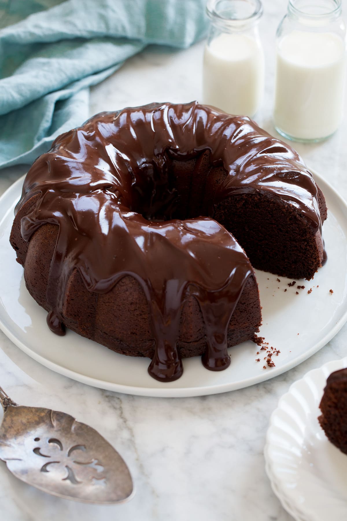 Chocolate Bundt Cake Whole chocolate bundt cake with one slice removed shown on a white platter with milk in the background and blue cloth.