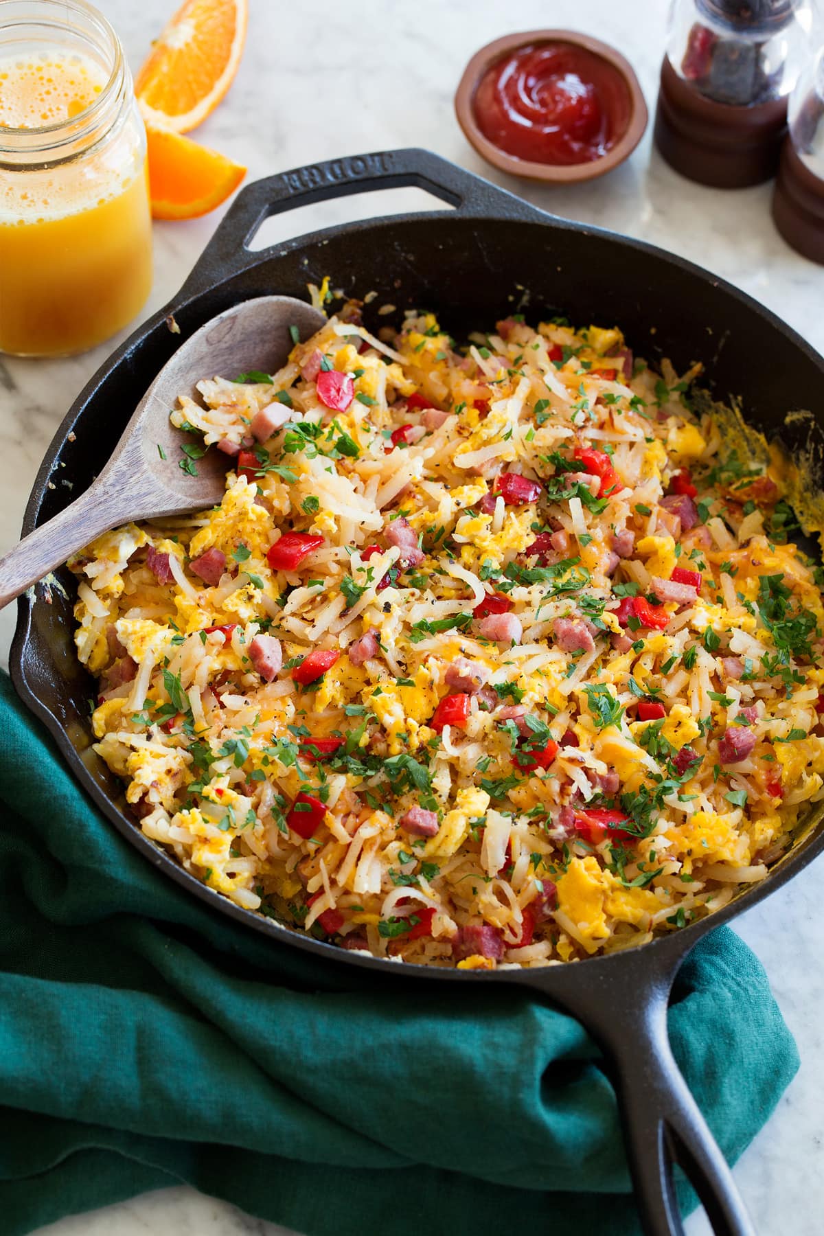 Breakfast Hash Photo: Egg and hash brown breakfast hash shown in a black cast iron skillet resting on a marble surface. A green cloth is shown to the side. Orange juice and ketchup are shown as serving suggestions.