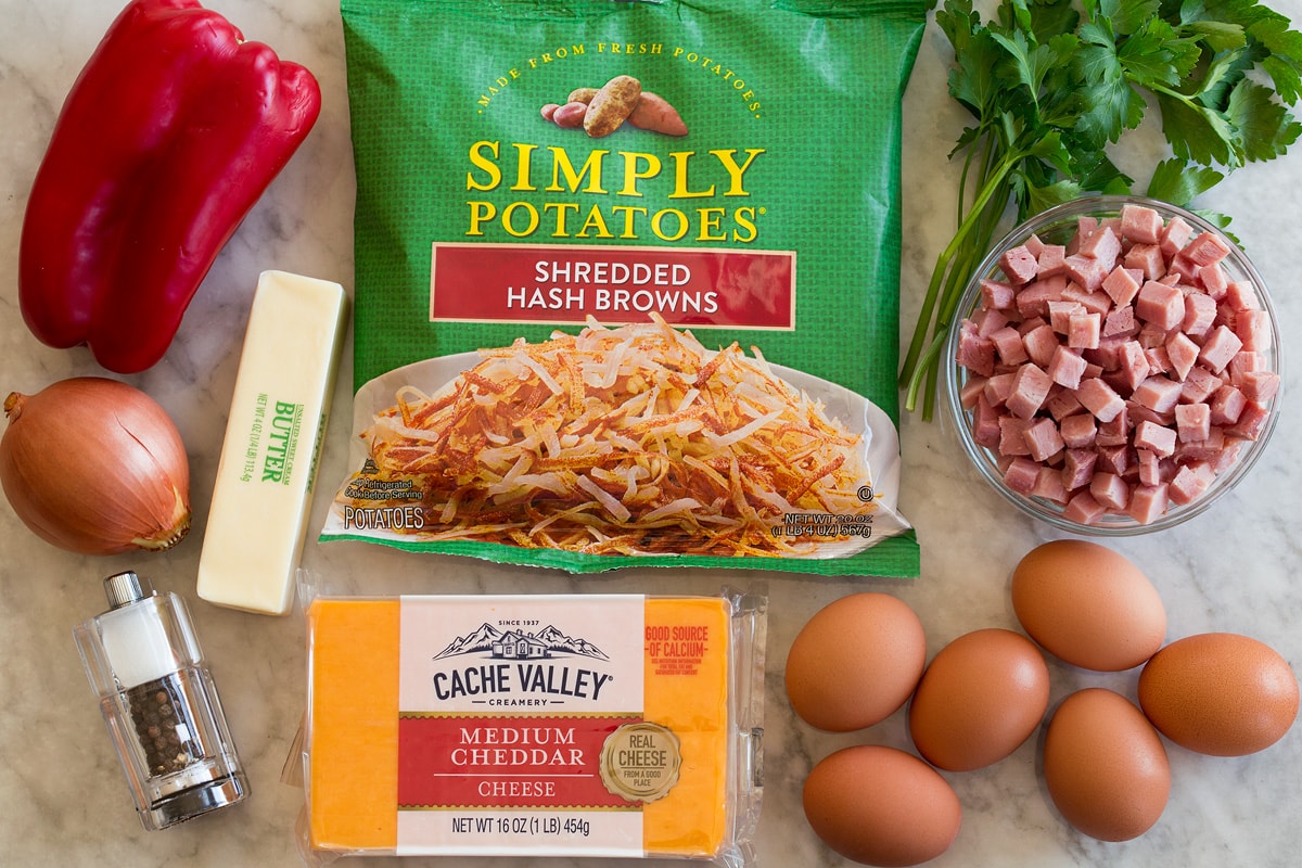 Breakfast Hash Photo: Ingredients shown that are used to make breakfast hash. Includes packaged shredded hash browns, bell pepper, cheddar cheese, eggs, butter, onion, ham, parsley, salt and pepper.