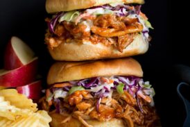 Photo: Two BBQ Pulled Chicken Sandwiches stacked on a wood platter. Apples slices and potato chips are shown to the side as serving suggestions.