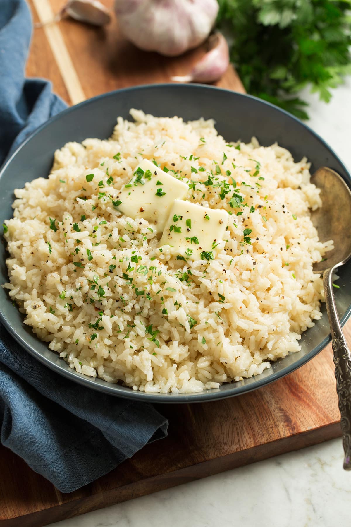 Garlic Butter Rice Rice seasoned with garlic and butter shown in a blue bowl over a wooden surface. Rice is topped with pieces of butter and chopped parsley.