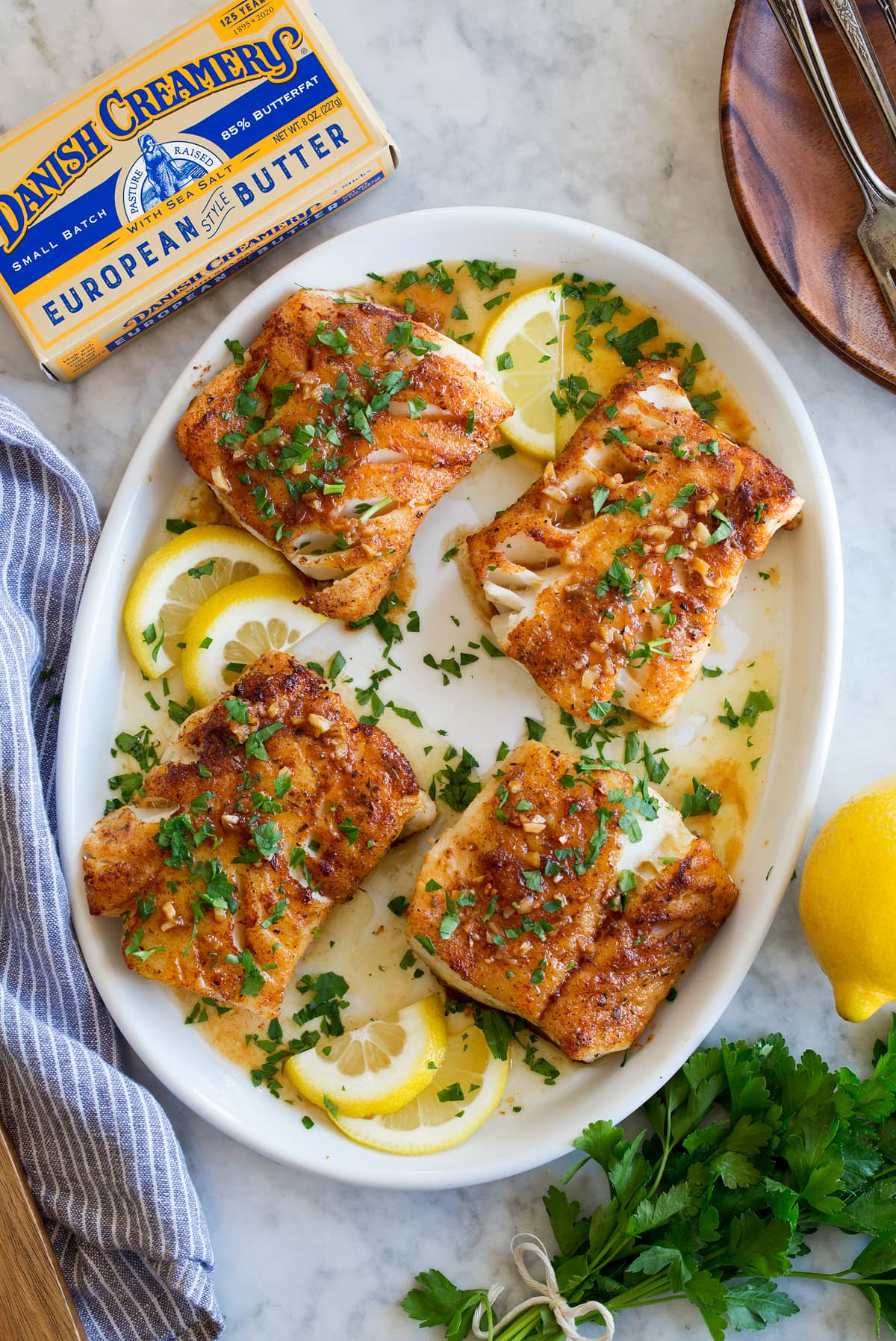 Lemon Butter Cod Recipe Four cod fillets shown overhead on a white oval platter. They are covered in a sauce and garnished with parsley. A package of butter and fresh parsley are shown to the side.